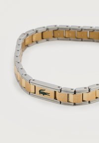 Δίχρωμο μεταλλικό βραχιόλι με ασημί άκρες και χρυσούς κρίκους, με μικρό εγχάρακτο λογότυπο κροκοδείλου σε έναν κρίκο.
