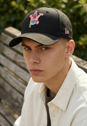 Junger Mann mit heller Haut, der eine schwarze Kappe mit einer bunten Cartoon-Figur trägt und auf einer Holzbank im Freien sitzt.