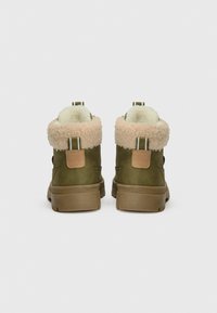 Grüne Knöchelschuhe mit einem flauschigen beigen Kragen, strukturiertem Stoff, kreisförmigem Absatz und kontrastierenden Hardware-Akzenten.