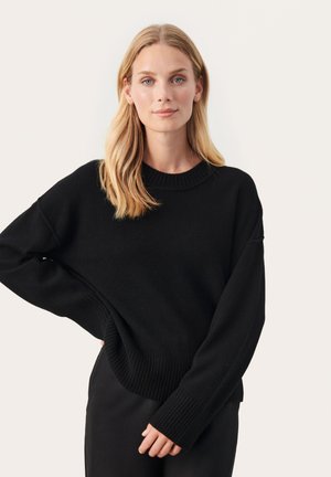 Svart stickad tröja med rund halsringning, sänkta axlar samt ribbade muddar och nederkant. Bärs över svarta byxor. Slät textur.