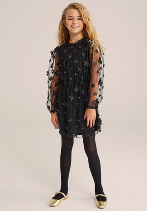Ragazza con lunghi capelli biondi che indossa un vestito in rete floreale nera, leggings neri e scarpe dorate, in piedi e sorridente su uno sfondo semplice.