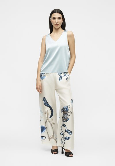 Femme portant un haut sans manches en satin bleu clair, un pantalon crème à jambes larges avec un imprimé floral et oiseau bleu, et des talons noirs ouverts à l'avant, debout avec une main dans la poche.