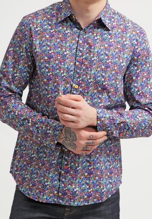 Mann trägt ein bunt gemustertes Hemd mit kreisförmigen Designs, die Hände gefaltet und zeigt Tattoos auf der linken Hand und den Fingern.