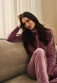 Fleecejakke med pink leopardprint, høj krave og lynlåslukning; parret med pink velourbukser, siddende på en beige corduroy-sofa.
