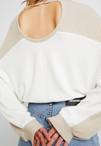 Un pull texturé crème et beige avec des manches larges et un dos ouvert, porté sur un jean bleu taille haute. Étiquette de marque visible à la taille.