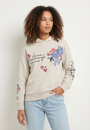 Woman with curly hair wearing beige hoodie with floral designs and "love always wins" text, paired with light blue jeans, standing casually.
