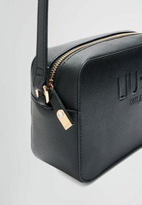 Sac synthétique noir texturé de forme rectangulaire, avec une fermeture éclair et des accessoires dorés. Présente un logo de marque embossé et une bandoulière réglable.