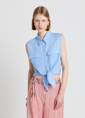 Jeune femme aux cheveux blonds courts portant une chemise sans manches bleu clair à boutons nouée à la taille et un pantalon rayé rouge et blanc avec des ornements.