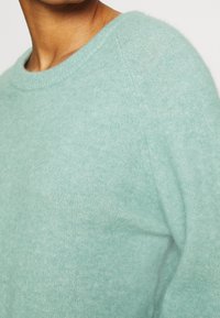 Gros plan d'une personne portant un pull en maille bleu clair doux avec un col rond sur un fond uni.