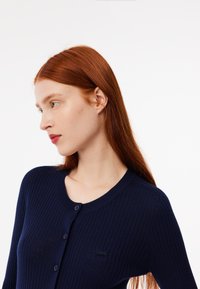 Cardigan a coste di colore blu navy con scollatura tonda, cinque bottoni e un piccolo logo sul lato sinistro. Presenta una silhouette aderente e un tessuto strutturato.