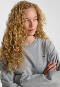 Donna bionda con capelli ricci che indossa una felpa grigia con un piccolo logo a forma di ape, braccia incrociate, che guarda di lato contro uno sfondo neutro.