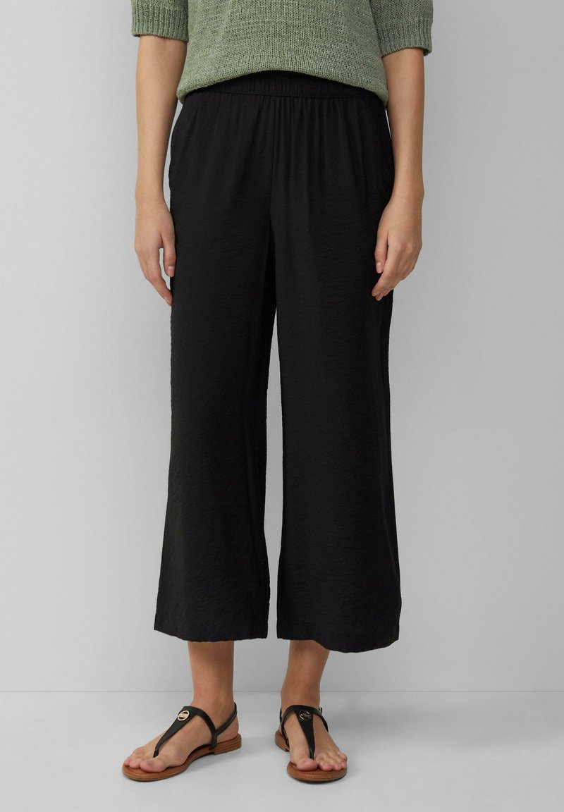 Femme portant un pantalon noir à jambes larges et cropped, un haut en tricot vert, et des sandales plates noires avec des semelles en bois, se tenant devant un fond uni.