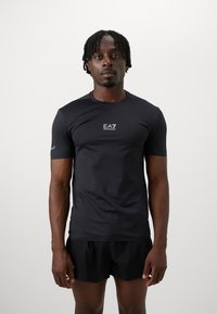 Svart t-skjorte med korte ermer laget av elastisk stoff, med en logo på brystet, sammen med svarte shorts.