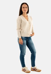 Femme portant un cardigan beige, un jean bleu et des baskets beiges avec des rayures bordeaux, se tenant avec une main dans la poche et souriant.
