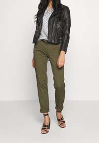 Black leather biker jacket with ribbed shoulder detail, patterned light blue blouse, olive green chinos, and strappy black high-heeled sandals.