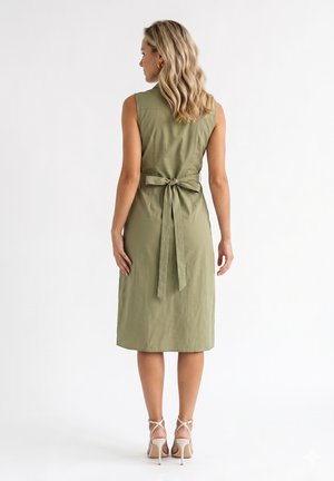 Femme aux cheveux blonds portant une robe midi sans manches vert olive nouée dans le dos avec un nœud, et des sandales beiges à talons hauts, debout dos à nous.