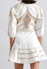 Robe blanche avec des détails en crochet complexes dans le dos et les manches, dotée d'une jupe évasée et de motifs texturés le long de l'ourlet.
