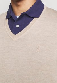 Pull beige à col en V superposé sur une chemise à col bleu marine, présentant un tricot texturé, un logo brodé et une finition en tissu lisse.