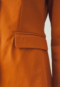 Blazer arancione con una texture liscia, dotato di una piccola tasca a pattina e una vestibilità sartoriale, che evidenzia linee pulite e cuciture ordinate.