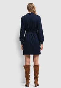 Granatowa sukienka kopertowa z długimi rękawami, przewiązana w talii, z guzikiem z tyłu, zestawiona z brązowymi zamszowymi botkami do kolan na obcasie.