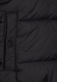 Svart boblejakke med quiltet tekstur, med en sirkulær svart knapp med "HUGO" og synlige sydetaljer. Lett materiale.