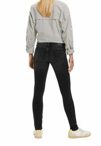 Femme aux cheveux blonds, portant un jean skinny noir, un tee-shirt à manches longues rayé et des baskets blanches, debout de dos.