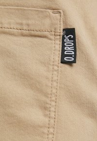 Beiger Stoff mit glatter Textur, ausgestattet mit einer Seitentasche und einem schwarzen Etikett mit weißem Text, auf dem "O.DROPS" steht.