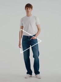 Mėlyni džinsai iš džinsinio audinio su tiesiomis kelnėmis, derinami su aptempta balta marškinėlių. Modelis stovi atsipalaidavęs, rankos kišenėse. Balti batai.