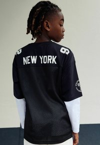 Patsient kahes konksus juustes vaatab külje poole, seljas kihiline must võrkkangas ja valge pikka varrukatega särk, mille seljal on "NEW YORK" tekst.