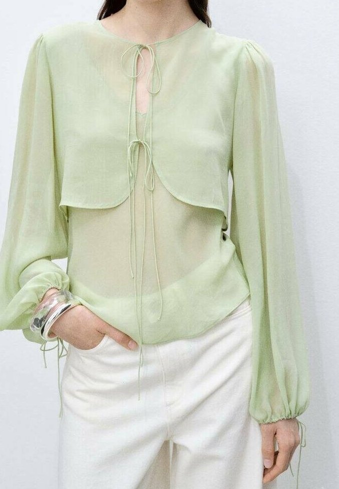 Femme portant un chemisier léger vert clair transparent avec de longues manches bouffantes et des liens à l'encolure, associé à un pantalon blanc taille haute.
