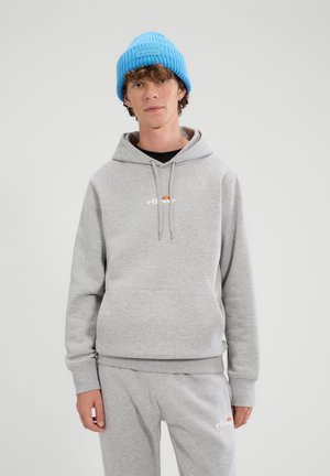 Jonge man draagt een lichtgrijze ellesse hoodie en joggingbroek met witte en oranje logo's, en een felblauwe gebreide muts tegen een effen achtergrond.