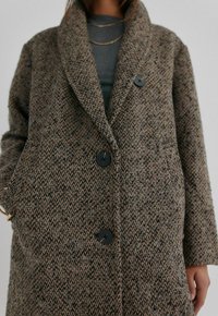 Brun vævet frakke med en tekstureret finish, oversized revers, to sorte knapper og sidelommer, båret over en grå top.