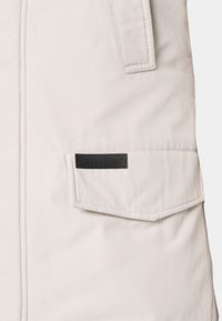 Giacca beige chiaro con un tessuto testurizzato, tasca a pattina con etichetta nera e dettagli cuciti lungo le cuciture.