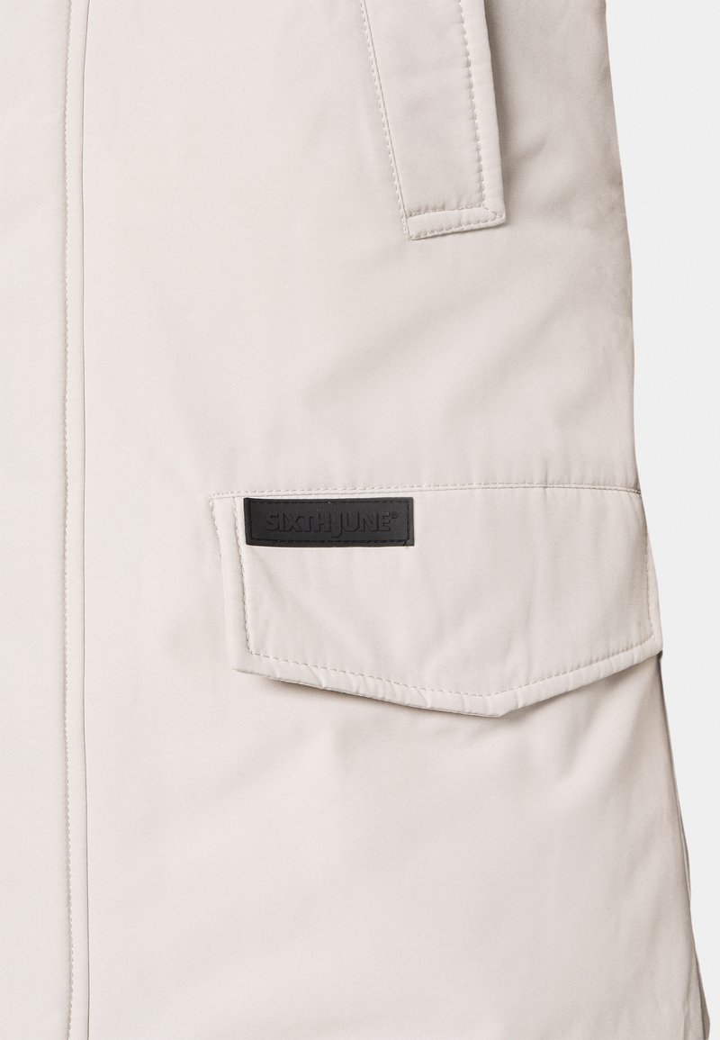 Veste beige clair avec un tissu texturé, une poche à rabat avec une étiquette noire et des détails cousus le long des coutures.