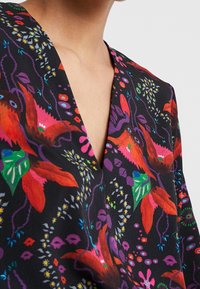 Chemisier à motif floral avec un décolleté en V profond, présentant des plantes rouges vives et vertes sur un fond noir, avec des accents subtils de violet et de bleu.