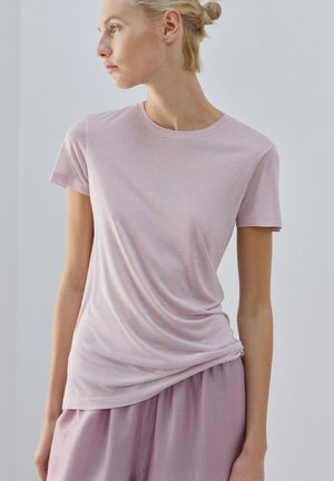 Młoda kobieta o blond włosach związanych do tyłu, ubrana w jasnoróżowy T-shirt z krótkim rękawem i pasujące luźne spodnie, spoglądająca w lewo na jednolitym tle.