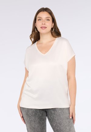 Cremfarbene Bluse mit V-Ausschnitt und kurzen Ärmeln, aus weichem, glattem Stoff. Kombiniert mit eng anliegenden grauen Jeans, die mit kleinen silbernen Nieten verziert sind.
