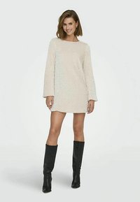 Donna sorridente che indossa un vestito mini a maniche lunghe di crema con texture e stivali neri fino al ginocchio, in piedi su uno sfondo chiaro semplice.