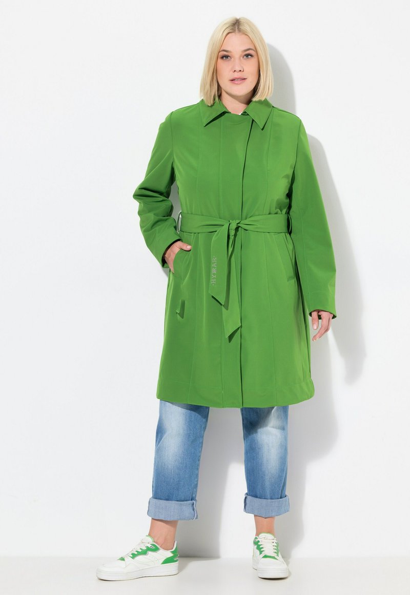 Cappotto trench verde con vita cintrata, maniche lunghe e due tasche frontali. Abbinato a jeans in denim blu arrotolati e sneakers bianche.