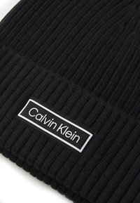 Sort strikket beanie med ribbet tekstur og et rektangulært hvidt mærke med "Calvin Klein"-logoet i sort skrift.