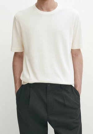 Mann trägt ein einfaches weißes kurzärmeliges T-Shirt, das in schwarze, hoch taillierte Hose gesteckt ist, mit den Händen in den Taschen.