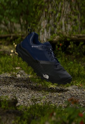 Sort og mørkeblå Gore-Tex vandresko med The North Face-logo, svævende over klippefyldt skovbund med grøn mos og træstammer i baggrunden.