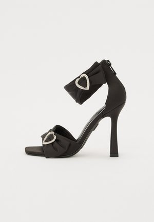 Mustad mustkõrgendusega sandaalid, millel on paela sõlm ja südamekujuline sädelev detail; tagaküljel on lukuga sulgemine ning sujuv, stiilne disain.