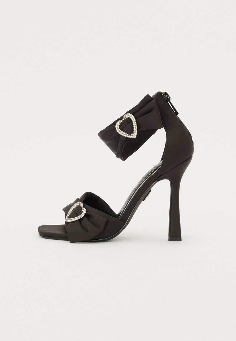 Melna augsta papēža sandale ar lentītes loku un sirds formas akmens akcentiem; ar rāvējslēdzēja aizdari aizmugurē un elegantu, gludu dizainu.