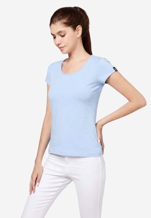 Frau mit braunen Haaren im Pferdeschwanz trägt ein hellblaues, eng anliegendes T-Shirt mit kurzen Ärmeln und weiße Hosen, steht mit einer Hand in der Hüfte.