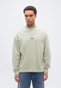 Άνδρας που φοράει ανοιχτό πράσινο φούτερ με μακριά μανίκια και μπλε λογότυπο "HUGO", συνδυασμένο με μπλε τζιν, στέκεται μπροστά σε απλό λευκό φόντο.