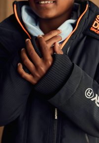 Giacca nera isolante con zip arancione, polsini a coste, logo sulla manica, indossata sopra un hoodie azzurro chiaro, mani incrociate all'altezza del petto.