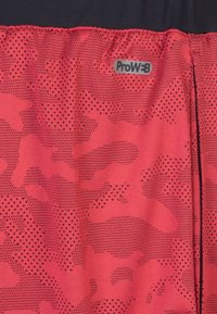 Tejido deportivo rojo con un patrón de camuflaje geométrico, que presenta texturas de puntos negros y el logo ProW+B en la cinturilla.