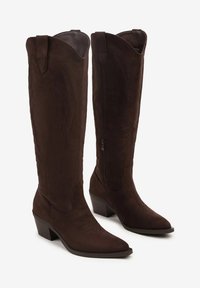 Bottes western en daim marron foncé, montantes jusqu'au genou, avec de petits talons carrés, bouts pointus, fermeture éclair sur le côté et broderies décoratives sur la tige et le pied.