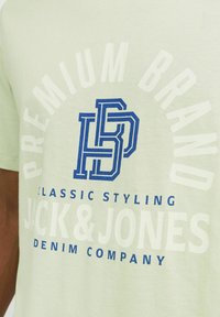 Gaiši zaļš kokvilnas T-krekls ar zilu grafiku, kurā rakstīts "PREMIUM BRAND" un "CLASSIC STYLING JACK & JONES DENIM COMPANY." Vienkāršs dizains.